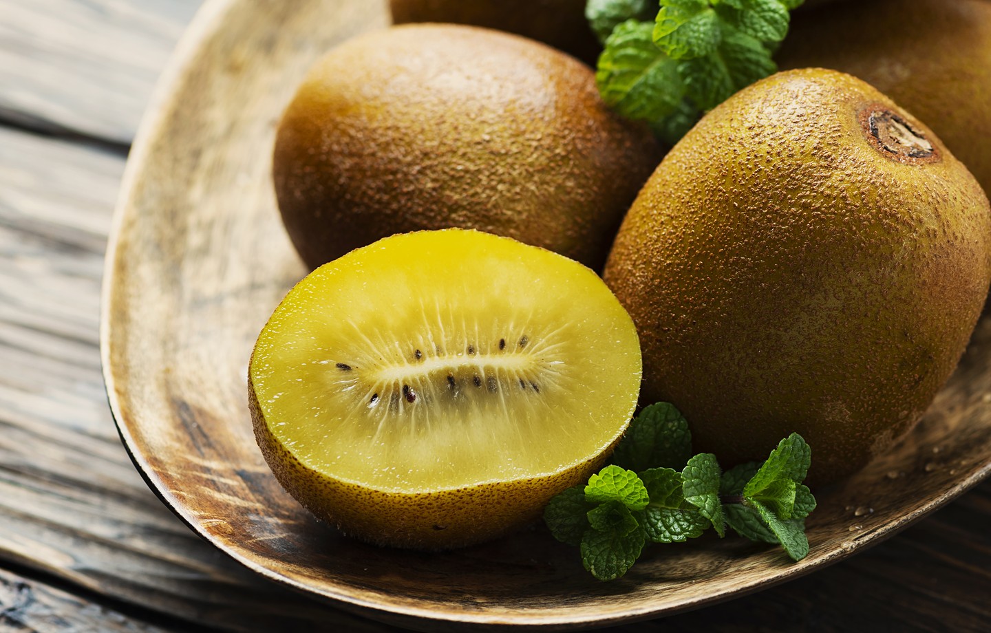 Kiwis golden entiers et un demi-kiwi à la chair jaune dans un plat en bois, avec des brins de menthe fraîche